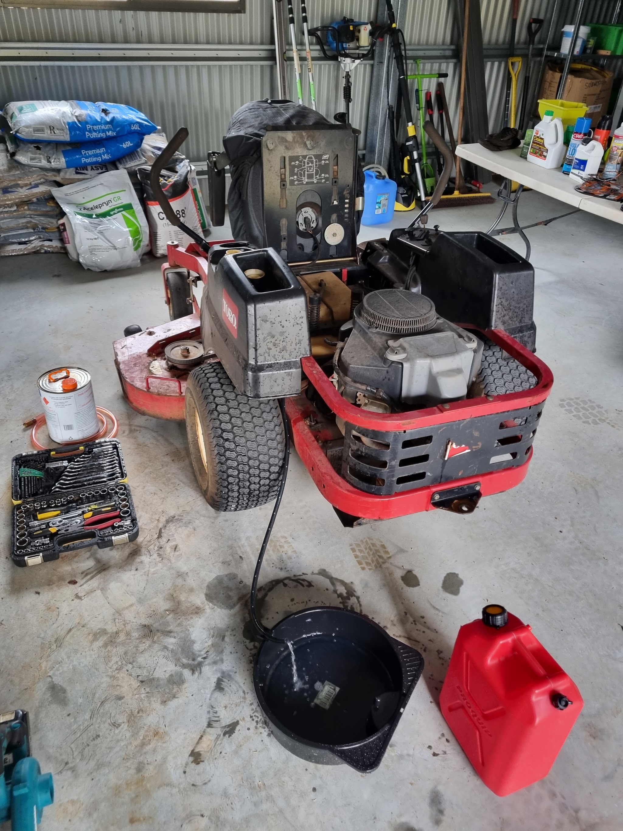 A zero-turn mower up on the workshop floor mid service, oil draining and tools laid out